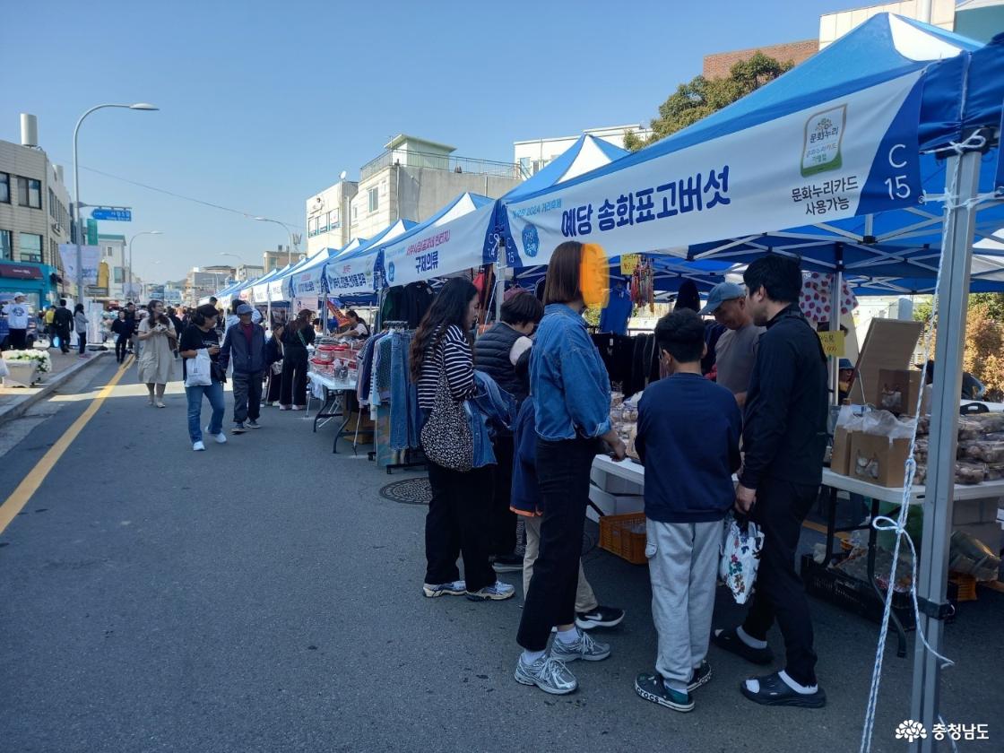 흰색 천막이 줄지어 설치되어 있고 각 천막마다 다양한 품목을 판매하는 상인들이 있으며 이를 구경하기 위해 많은 주민들이 오가며 보고 있다.