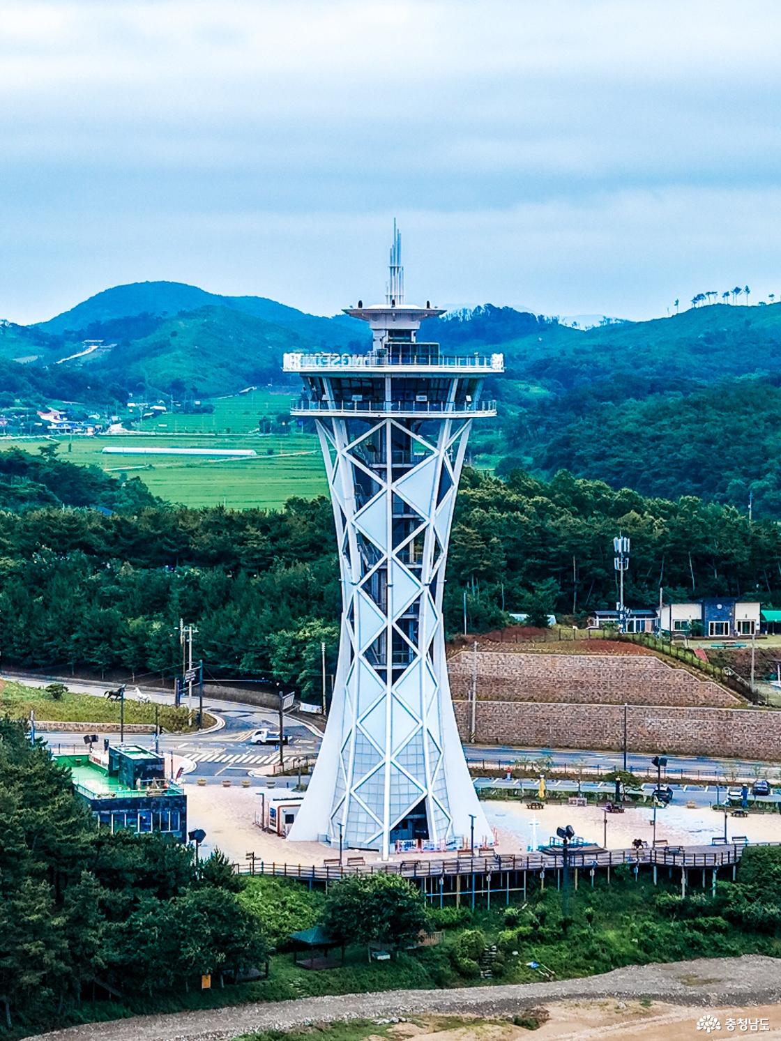 울창한 초록 산을 배경으로 흰색 X자 격자 구조의 홍성 스카이타워가 하늘을 향해 솟아 있고, 타워 하단에 주차장과 부대시설이 갖춰져 있다.