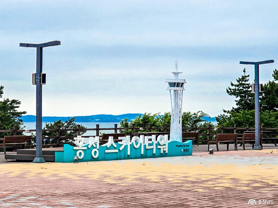 타워 입구 광장에 청록색 바탕에 흰 글씨로 '홍성 스카이타워' 조형물이 놓여 있고, 그 뒤로 타워 상부와 서해 바다가 배경으로 펼쳐져 있다.